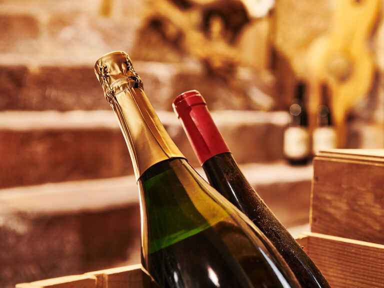 Zwei Sommeliers, Roland Berlin und Michael Kolb, garantieren eine exzellente Weinauswahl und runden damit jedes Gericht des Küchenteams ab. Eine Flasche Champagner und eine Flasche Wein ragen aus einer Holzkiste, die sich in einem Gewölbekeller befindet.