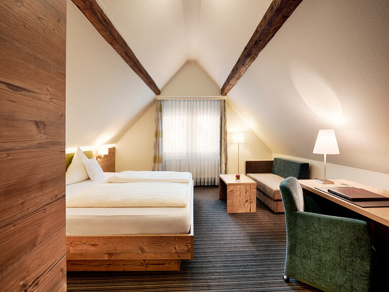 Die Hotelzimmer im Stammhaus Hotel Kronelamm sind aufgrund des gemütlichen Landhausstils sehr beliebt. Ein Doppelbett, eine Couch, Schreibtisch sowie ein grüner Sessel stehen in einem Zimmer mit Dachschräge.