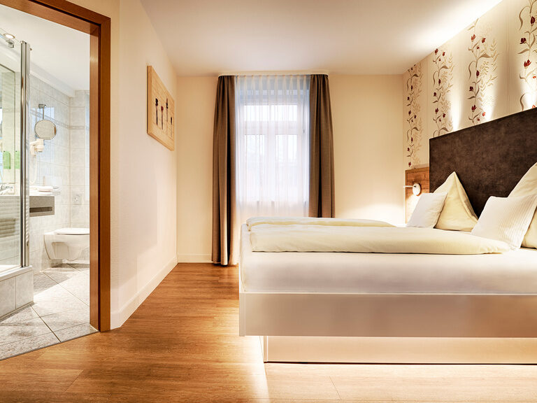 Ein großzügiges Doppelzimmer mit Doppelbett, welches mit Lichtern beleuchtet wird, und Zugang zum Badezimmer mit Dusche.