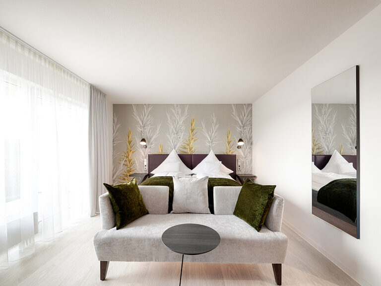 Die Zimmer mit gemütlicher Sitzgelegenheit liegen im zweiten Stock des familiengeführten Hotels Kronelamm im Schwarzwald. Vor einem Doppelbett, auf dem weiße Bettwäsche und grüne Bettaccessoires angerichtet wurden, steht eine hellgraue Couch.